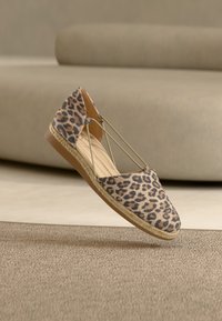 Ravni čevelj espadrille z leopardjim vzorcem, bež elastiki in tkanim podplatom iz juta na bež preprogi in ozadju kavča.