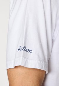 Hvit bomull T-skjorte med ermer som har marinblå kursiv broderi med ordet "malbon." Teksturen ser myk og glatt ut.