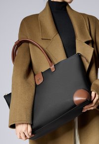 Osoba oblečená v kamelovém kabátu a černém topu drží velkou černou tašku s hnědými koženými uchy a detaily.