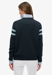 Marineblauer Pullover mit hellblauen Streifen an den Ärmeln, strukturierter Strick, gerippter Saum, getragen über einem gestreiften Hemd mit Kragen, kombiniert mit weißen Hosen.