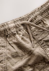 Pantaloncini mimetici beige con vita elastica e chiusura a coulisse. La trama del tessuto sembra leggera con motivi delicati.