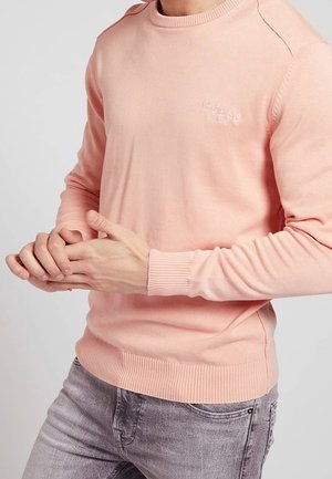 Man wearing light pink Guess sweater with ribbed cuffs and hem, paired with faded gray jeans, hands clasped in front.
