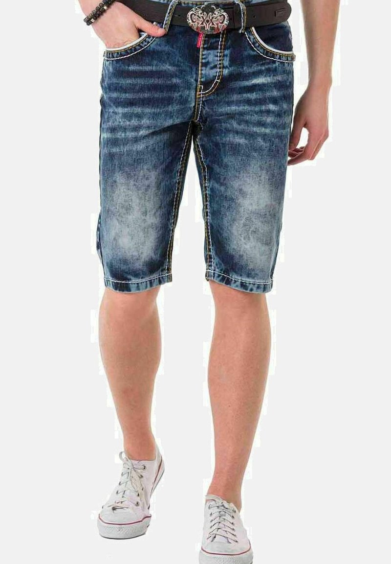 Jeansshorts in einer verblassten blauen Waschung mit kontrastierenden gelben Nähten, fünf Taschen-Design und einer rohen Kante, getragen mit weißen Sneakern.