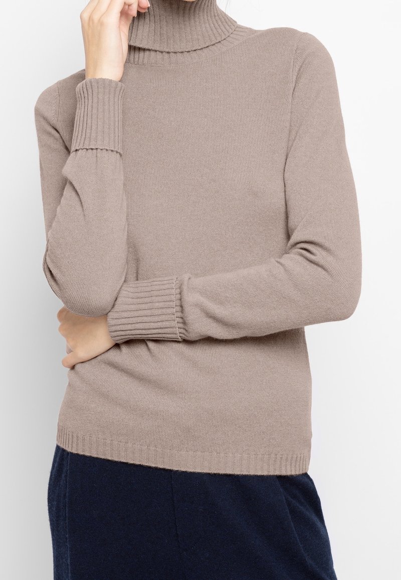 Personne portant un pull à col roulé côtelé couleur taupe avec les manches couvrant partiellement les mains, associé à un pantalon bleu marine foncé.