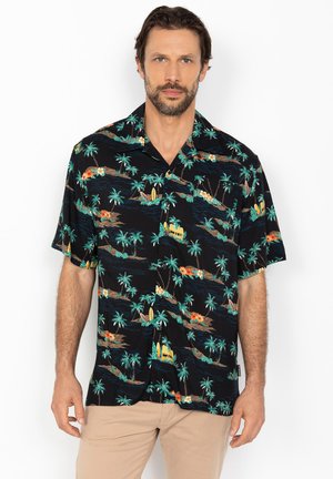 Mężczyzna w czarnej hawajskiej koszuli z wzorami palm, desek surfingowych i wysp, w beżowych spodniach, stojący na białym tle.