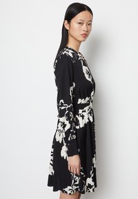 Frauen mit glattem schwarzen Haar, die ein schwarzes Kleid mit großen weißen Blumenmustern tragen, stehen seitlich vor einem schlichten weißen Hintergrund.