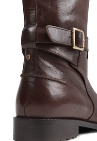Botte en cuir marron hauteur genou avec détail de boucle en or, texture lisse et talon en bois empilé.