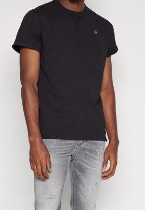 Mann trägt ein schlichtes schwarzes Kurzarm-T-Shirt mit kleinem Logo auf der Brust und hellgraue, abgenutzte Jeans vor weißem Hintergrund.