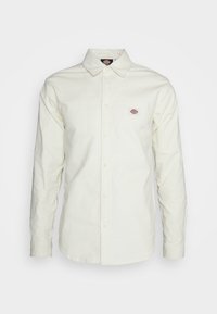 Camisa de botones color crema claro con cuello clásico, que presenta un pequeño parche de logo en el pecho izquierdo y mangas largas. Tejido de algodón suave.