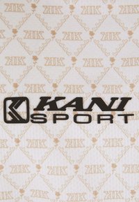 Logo KANI SPORT černé barvy na světlém textilním pozadí s opakujícím se vzorem stylizovaných srdcí a písmen "K" v béžové barvě.