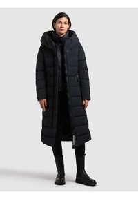 Lungo cappotto imbottito nero con cappuccio, chiusura con zip, tasche laterali e un design a quadri rettangolari. Indossato sopra leggings scuri e stivali.