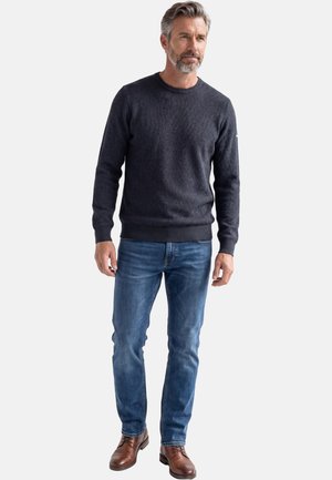 Man met grijs haar en baard, gekleed in een donkergrijze trui, blauwe jeans en bruine leren schoenen, staande tegen een effen lichte achtergrond.