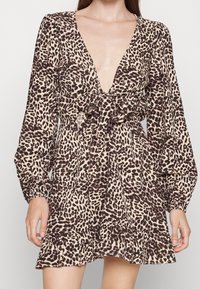 Nainen, jolla on yllään pitkähihaisen, syvällä V-kaula-aukolla varustettu leopardikuvioinen mekko, jossa on edessä satiininen solmu ja röyhelöhelma, seisoo tasaisella taustalla.