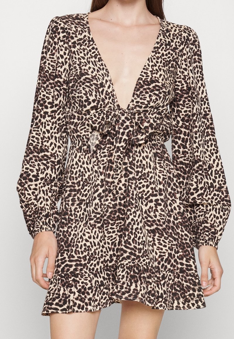 Nainen, jolla on yllään pitkähihaisen, syvällä V-kaula-aukolla varustettu leopardikuvioinen mekko, jossa on edessä satiininen solmu ja röyhelöhelma, seisoo tasaisella taustalla.