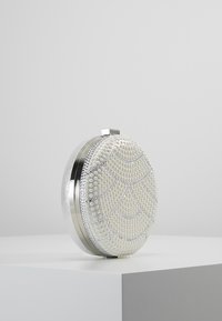 Biała owalna kopertówka o koralikowej fakturze, ze srebrnymi akcentami i okrągłym wzorem. Wyposażona w metalowe zapięcie i błyszczące wykończenie.