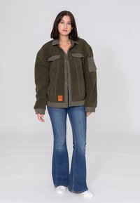 Zielony polarowy kurtka w kolorze oliwkowym z zapięciem na guziki, kieszeniami na piersi i kieszenią na suwak na rękawie; połączona z dzwonami w kolorze niebieskim i białymi trampkami.