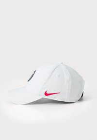 Biela baseballová čiapka s ohnutým okrajom, na prednej strane má námornícku logovú nášivku a na boku červený Nike swoosh.