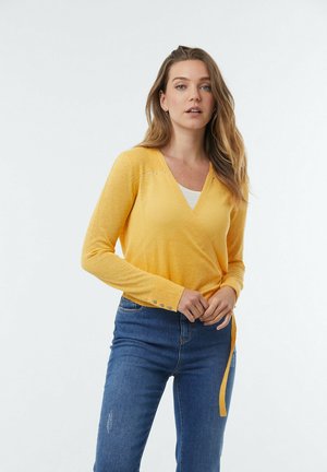 Cardigan jaune en matière douce et légère, avec des manches longues et des embellissements subtils près de l'encolure. Associé à un jean bleu.