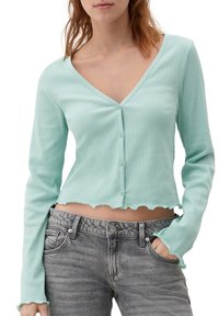 Mátově zelený vroubkovaný kardigan s dlouhým rukávem, V-výstřihem, zkráceným střihem, scalloped lemem a ladícími knoflíky. Nosený s šedými denimovými džíny.