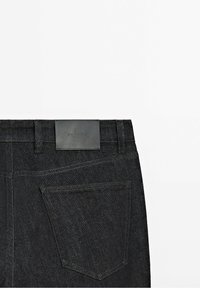 Vue de dos d'un jean en denim noir montrant la poche arrière droite et une étiquette de marque en cuir noir au-dessus de la poche sur la ceinture.