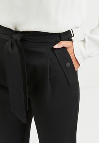 Zwarte broek met een gestrikt middel, gemaakt van een gladde stof, met voorzakken en een knoopdetails op de zakflap.