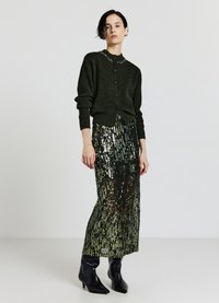 Cardigan vert foncé à encolure ronde et fermeture à boutons, associé à une jupe longue pailletée brillante aux tons verts. Bottines noires.