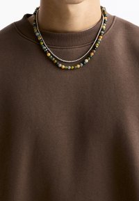 Brun sweatshirt med ribbet halsudskæring, lagdelt med to halskæder: en sølvkæde og en perlekæde med flerfarvede sten.