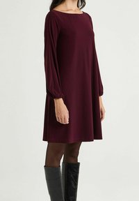 Robe trapèze bordeaux avec manches longues bouffantes et encolure bateau, fabriquée dans un tissu léger ; portée avec des bottes noires montantes jusqu'aux genoux.
