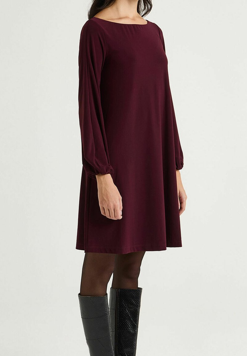 Robe trapèze bordeaux avec manches longues bouffantes et encolure bateau, fabriquée dans un tissu léger ; portée avec des bottes noires montantes jusqu'aux genoux.