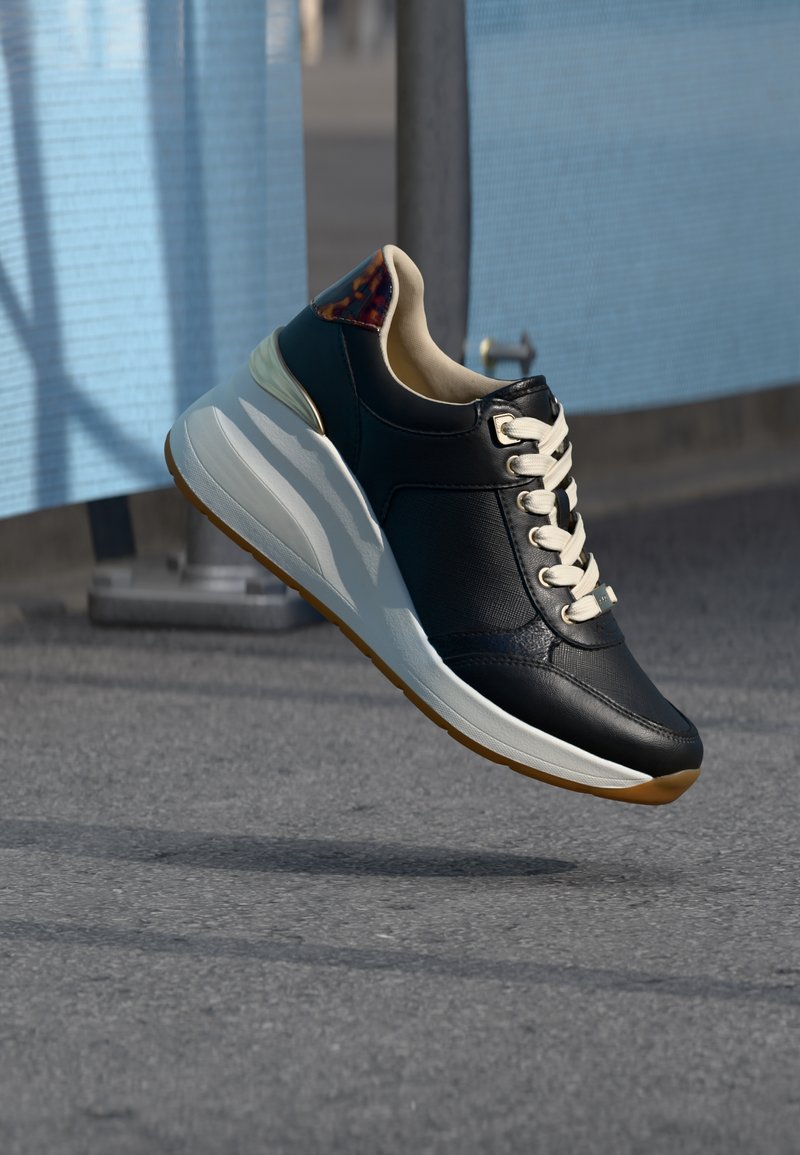 Baskets noires avec semelle blanche et lacets flottant au-dessus d'un trottoir gris, tissu bleu et poteau en métal en arrière-plan flou.