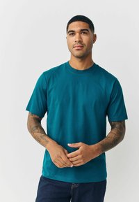 Teal krāsas kokvilnas T-krekls ar apaļu kakla izgriezumu, īsām piedurknēm un brīvu piegriezumu. Gluda tekstūra un vienkāršs dizains, apvienots ar tumšām džinsām.