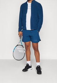 Sininen vetoketjullinen takki valkoisen paidan päällä, siniset shortsit, joissa on sana "BORG", ja mustat urheilukengät. Pitelee tennismailaa.