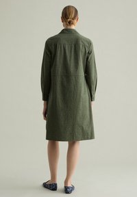 Olivgrünes, knielanges Hemdkleid mit Kragen, langen Ärmeln und feinem strukturierten Muster. Tragen mit dunkelblauen flachen Schuhen.