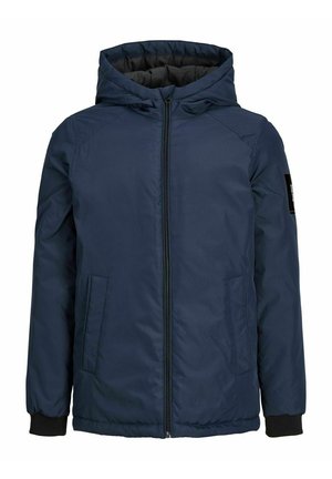 Jeune personne portant une veste bleu marine, un t-shirt blanc graphique, un jean bleu et des baskets blanches, debout devant un fond uni.