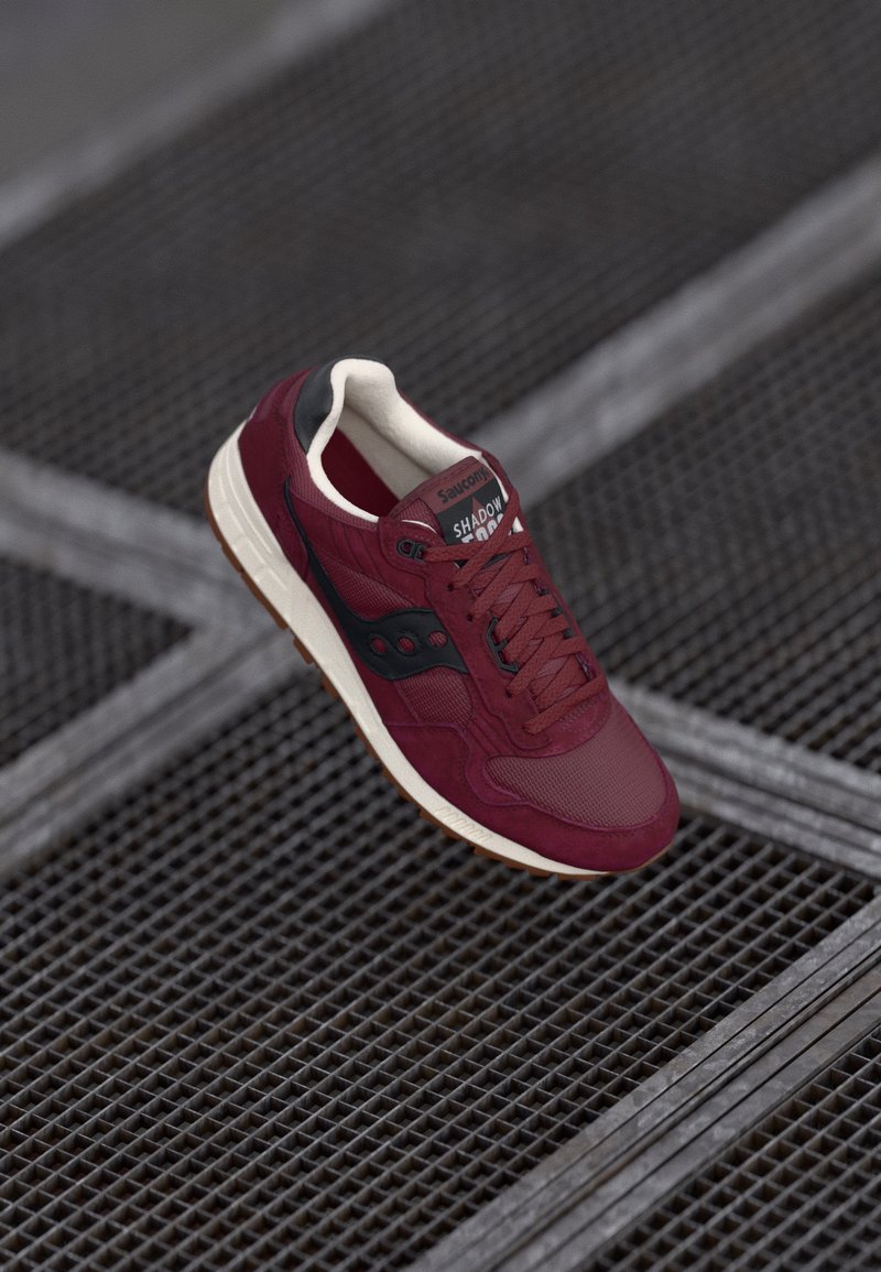 Burgunderfargede sneakers laget av semsket skinn og mesh, med svarte detaljer, hvit såle, avrundet tå, og teksturert overflate med synlige snører.