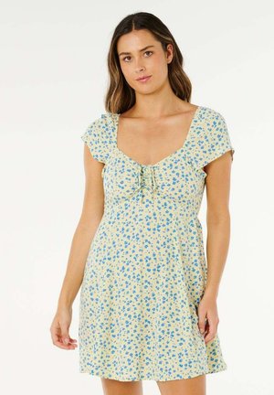 Femme aux cheveux bruns portant une robe jaune à manches courtes avec un motif floral bleu et un col carré, debout devant un fond blanc.