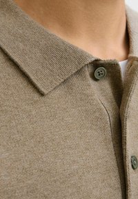 Kratak pogled na bež pleteni ovratnik polo majice s dugmadi, nosi se preko bijele majice na golu kožu.