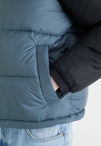 Blauwe en zwarte pufferjas met een gequiltde textuur, voorzien van een zijzak en elastische manchetten, gemaakt van waterafstotend materiaal.