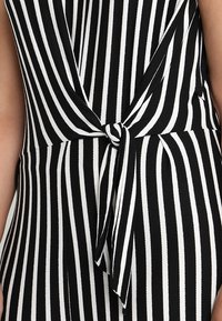 Robe portefeuille à rayures verticales noires et blanches avec un nœud à l'avant à la taille, montrant une partie du bras gauche.