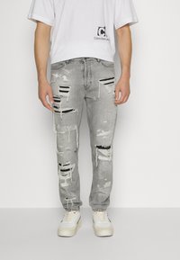 Mies, joka käyttää vaaleanharmaita kuluneita farkkuja mustilla kangaspaloilla, valkoisia tennareita ja valkoista Calvin Klein -t-paitaa, seisoo valkoista taustaa vasten.