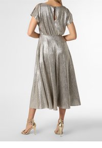 Femme portant une robe midi métallisée argentée à manches courtes et dos en découpe, associée à des sandales à talons hauts dorées avec des lanières.