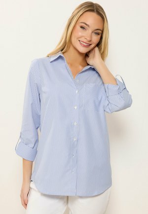 Smilende blond kvinde iført en lyseblå og hvid, vertikalt striped skjorte med op-rullede ærmer og hvide bukser.