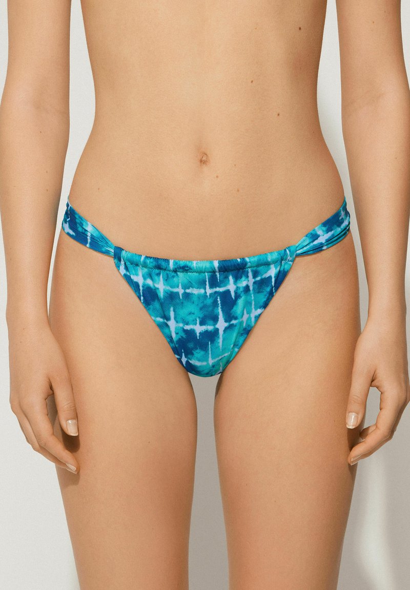 Bikini spodní část s batikovaným vzorem v odstínech modré a tyrkysové. Obsahuje měkkou látku, nízký střih a uzlové boční pásky s hladkým povrchem.