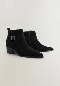 Botas de tobillo de ante negras con punta afilada, tacón cuadrado y cremallera lateral. Presenta una correa decorativa con un acento de hebilla plateada.
