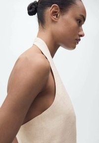 Beige halter neck top made of textured fabric, featuring wide armholes and a relaxed fit, shown from the side against a neutral background.