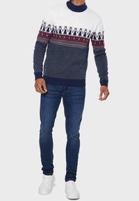 Pull en tricot navy et blanc avec des motifs décoratifs rouges et blancs, doté d'un col rond et de poignets côtelés, associé à un jean foncé et des baskets blanches.