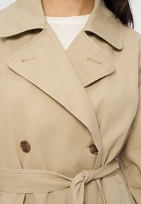 Trench-coat beige croisé à larges revers, deux boutons marron visibles, et une ceinture nouée à la taille sur un haut blanc.