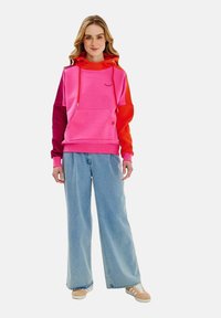 Světle růžová mikina s barevně blokovanými rukávy v odstínech růžové a červené. Obsahuje klokaní kapsu a kapuci se šňůrkou. V kombinaci s kalhotami s širokými nohavicemi.