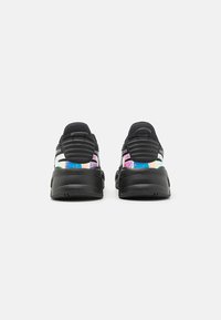 Ein Paar schwarze Sportschuhe, die von hinten mit dicken Sohlen und schillernden Elementen über den Fersen vor einem schlichten weißen Hintergrund gezeigt werden.