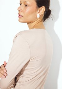 Haut rose pâle à manches longues en tissu texturé, au design ajusté et avec de discrètes fronces à l'épaule, associé à des boucles d'oreilles blanches accrocheuses.